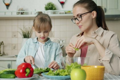 Cute loving daughter is helping mother with salad cutting cucumbers feeding young woman and laughing having fun at home. People and cookery concept.