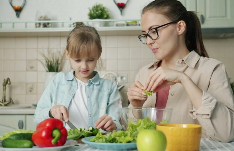 Cute loving daughter is helping mother with salad cutting cucumbers feeding young woman and laughing having fun at home. People and cookery concept.