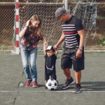 Full body of positive happy parents in casual clothes holding toddler son hands while walking together on football field on sunny day