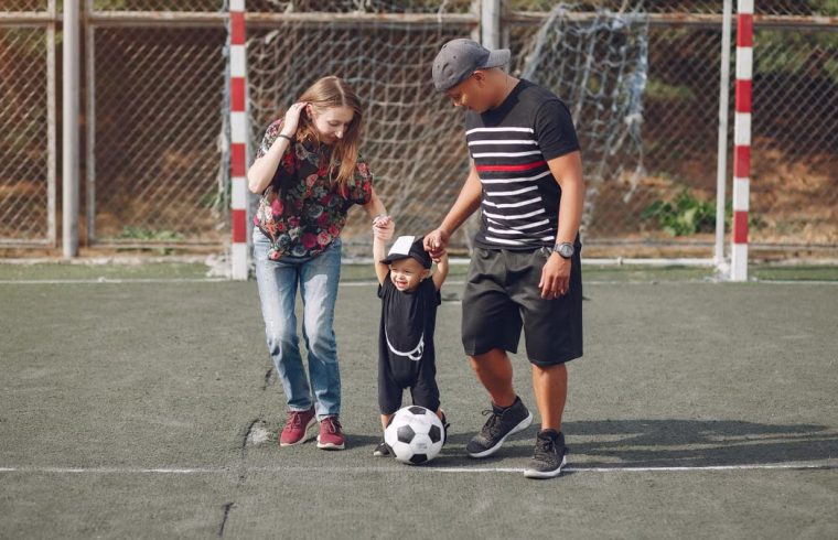 Full body of positive happy parents in casual clothes holding toddler son hands while walking together on football field on sunny day